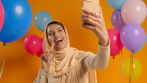 Retrato-De-Estudio-De-Una-Mujer-Tomándose-Un-Selfie-Usando-Hijab-Celebrando-Una-Fiesta-De-Cumpleaños-Rodeada-De-Globos
